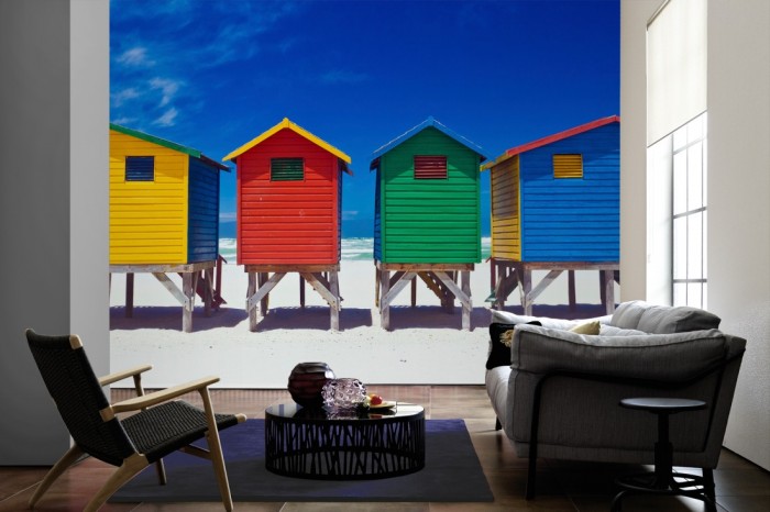 Im Wohnzimmer die Fototapete Bunte Strandhäuser in Südafrika, Muizenberg auf einer großen Wand tapeziert. Die Fototapete Bunte Strandhäuser in Südafrika, Muizenberg ist als Vlies-Fototapete im Wohnzimmer leicht zu tapezieren.