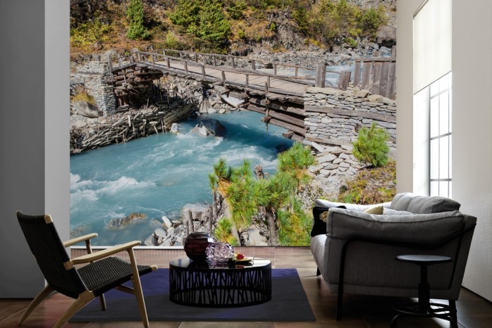 Im Wohnzimmer die Fototapete Bach mit Holzbrücke im Gebirge auf einer großen Wand tapeziert. Die Fototapete Bach mit Holzbrücke im Gebirge ist als Vlies-Fototapete im Wohnzimmer leicht zu tapezieren.
